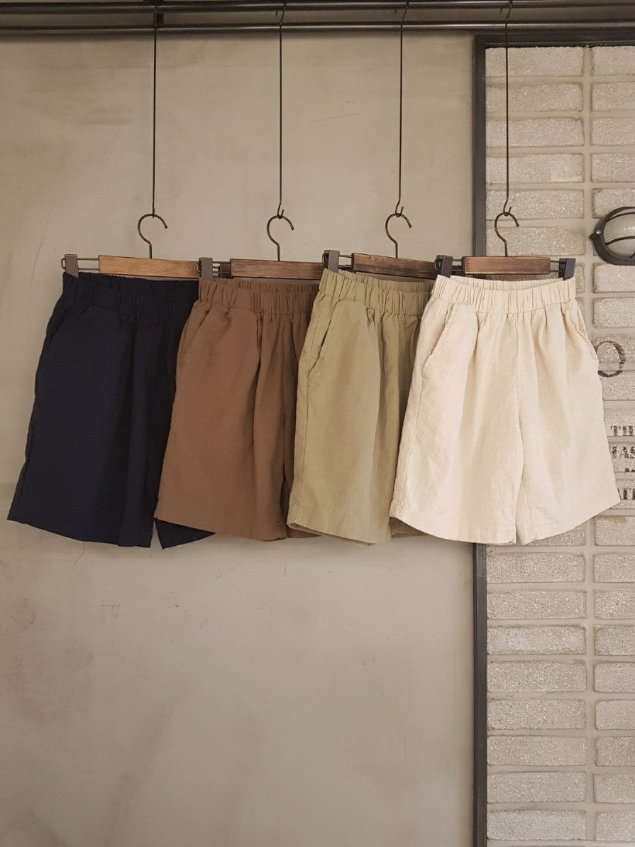 Four pairs of linen shorts hanging on wooden hangers. The shorts are in navy blue, brown, light green, and cream colors. The shorts have an elastic waistband and are made of a lightweight fabric.
